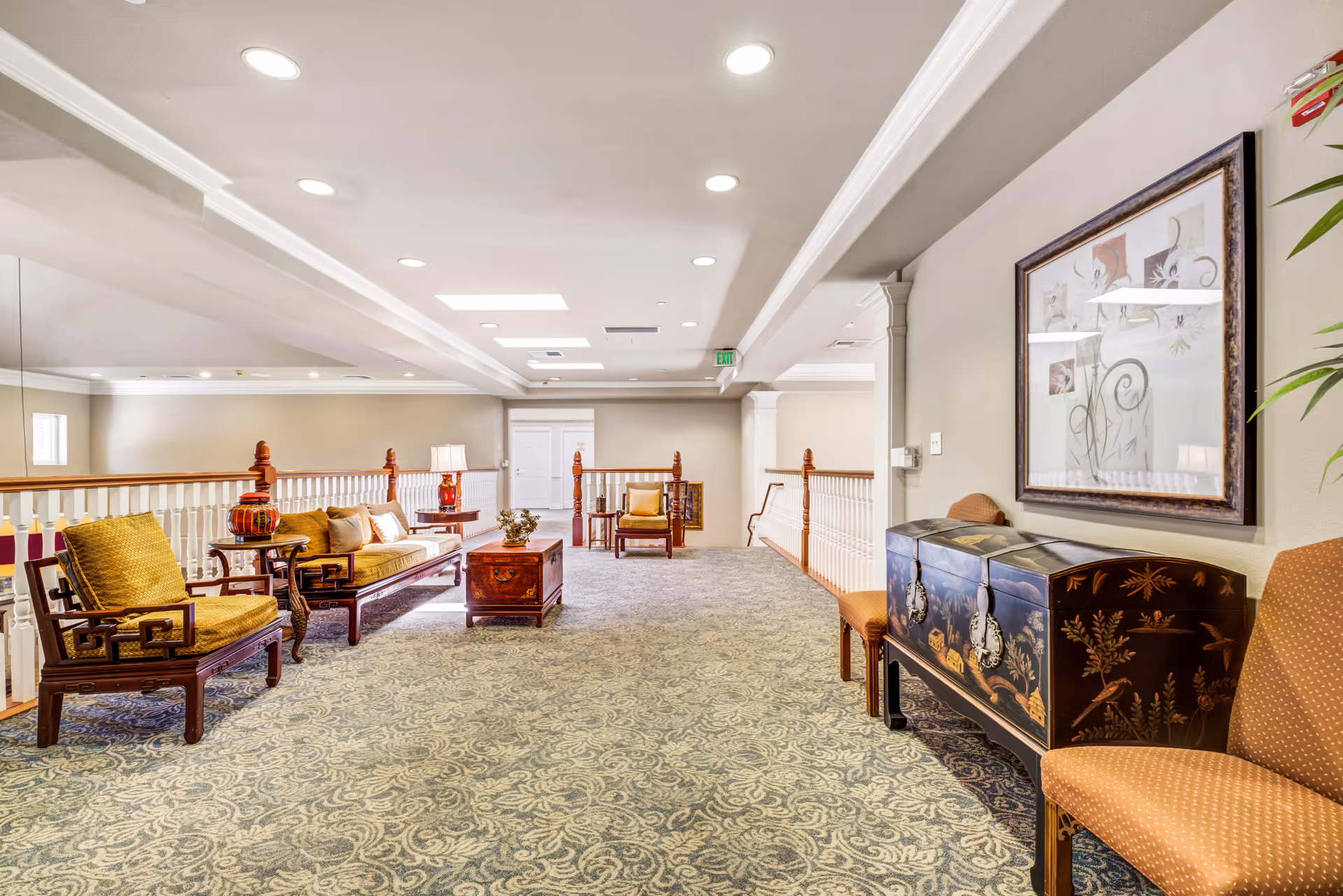 A spacious, well-lit common area with patterned carpet flooring, wooden furniture including chairs and a sofa with cushions, a wooden chest as a coffee table, and decorative items such as a framed artwork on the wall and a potted plant. The area has white railings and recessed ceiling lights.