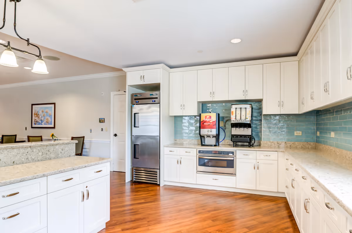 A bright and spacious kitchen area with white cabinetry, a stainless steel refrigerator, built-in microwave, and coffee and beverage dispensers on the countertop. The backsplash features light blue tiles, and the floor is a warm wood tone. In the background, there is a dining area with chairs and a framed picture on the wall.