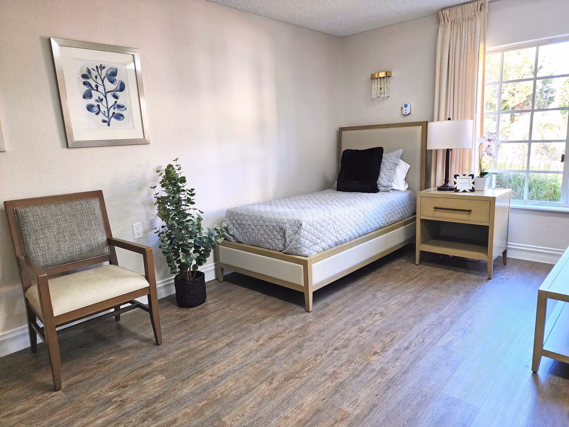 A bright and clean bedroom with a single bed featuring a gray quilted bedspread and multiple pillows. Next to the bed is a wooden nightstand with a lamp, a small plant, and a decorative clock. A wooden chair with cushioned seat and backrest is positioned near a potted plant. The room has light-colored walls, wood flooring, a large window with curtains letting in natural light, and framed botanical artwork on the wall.