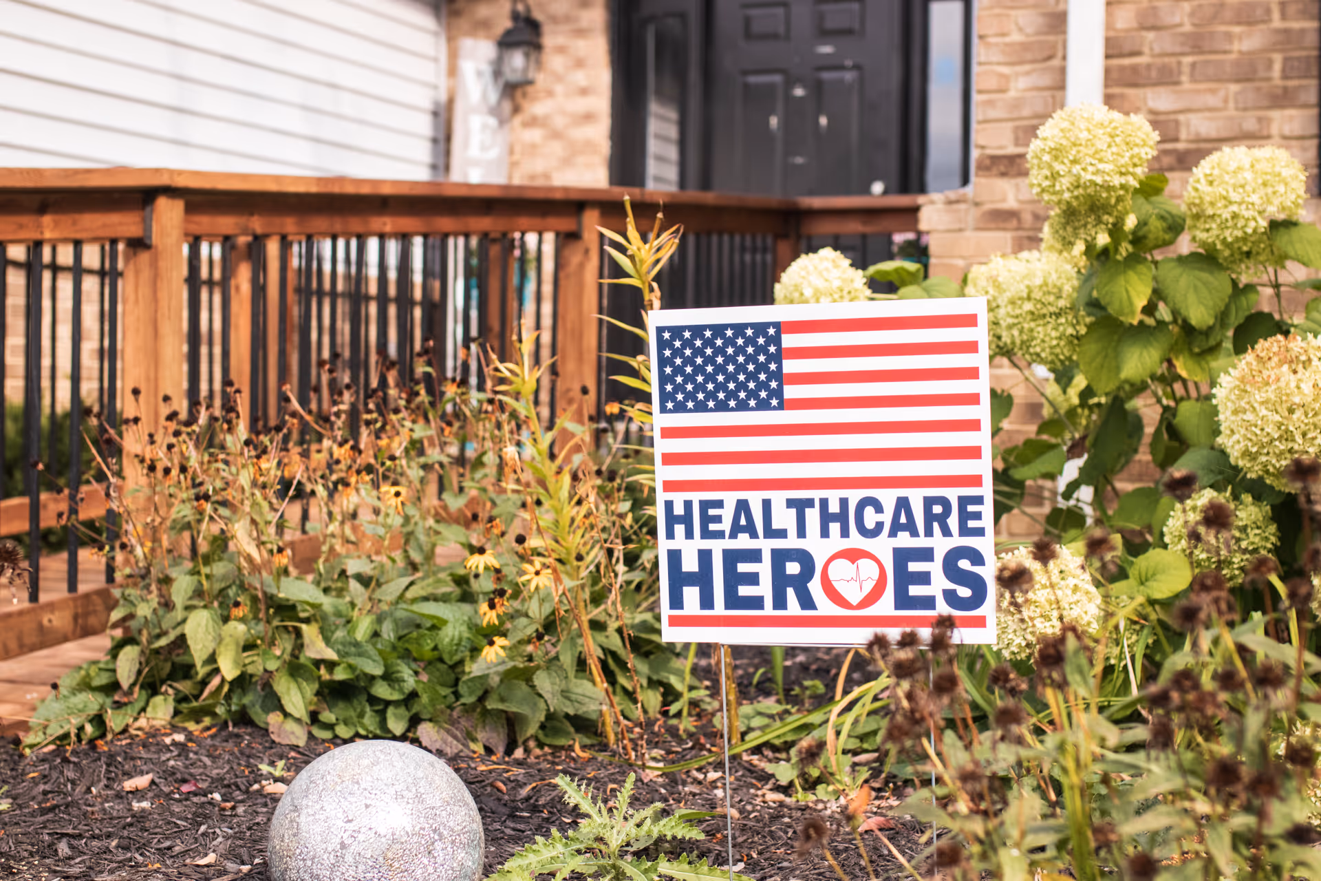 A garden area in front of a building entrance with a wooden railing and various plants. A sign with an American flag design reads 'Healthcare Heroes' with a heart and heartbeat line graphic.
