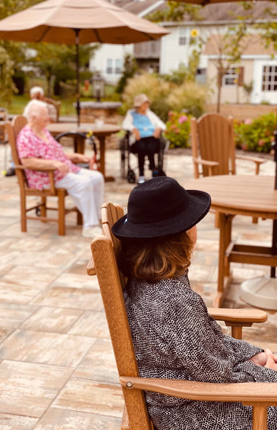 Several elderly individuals sitting outdoors on wooden chairs around tables on a tiled patio. One person in the foreground is wearing a black hat and patterned shirt, while others are seated in the background near an umbrella and greenery.