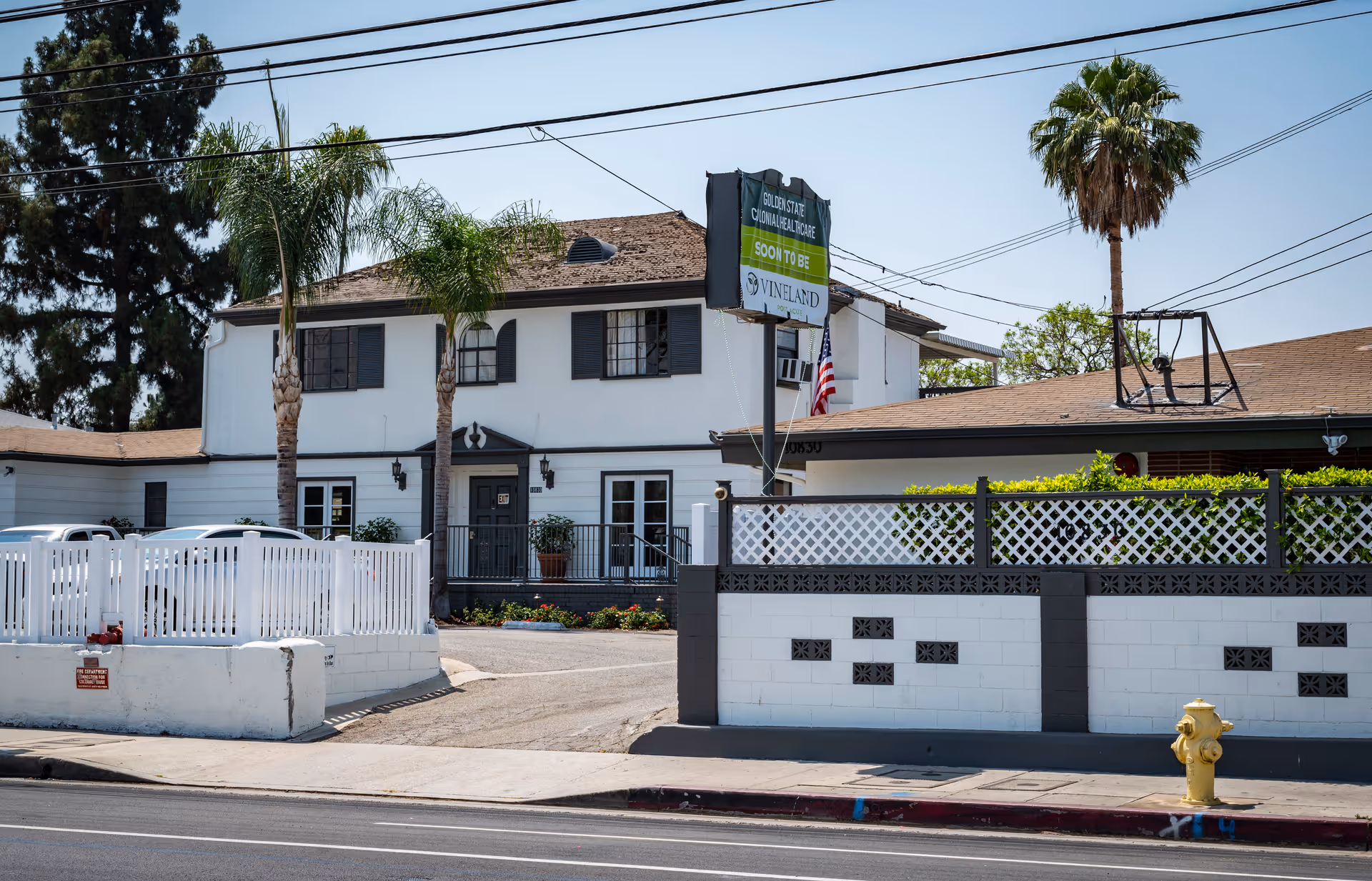 Exterior view of a two-story white building with black shutters and a brown roof, surrounded by a white fence and some palm trees. A sign in front reads 'Golden State Convalescent Home Soon to Be Vineland'. There is a yellow fire hydrant on the sidewalk and an American flag near the building entrance.