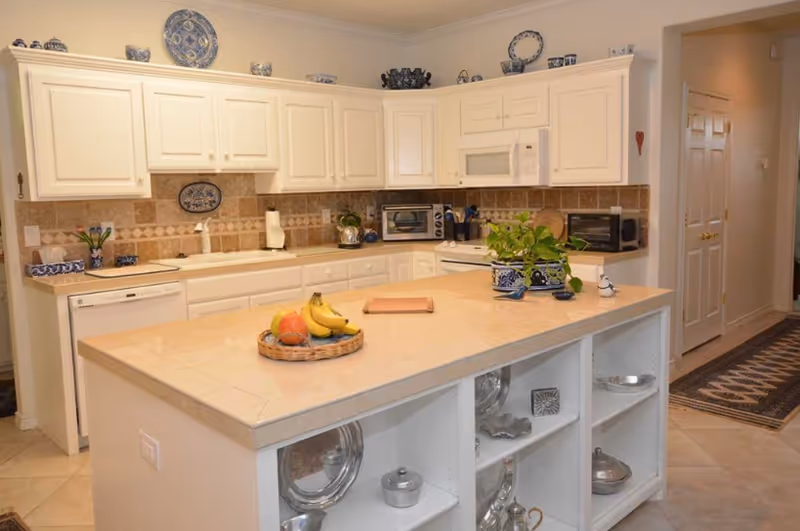 A bright kitchen with white cabinets and beige tiled countertops and backsplash. The kitchen features a large island with open shelving displaying silver dishes and containers. On the island countertop, there is a basket with fruit and a potted plant in a blue and white ceramic pot. The kitchen includes appliances such as a dishwasher, microwave, toaster oven, and stove. Decorative blue and white ceramics are placed on top of the cabinets.