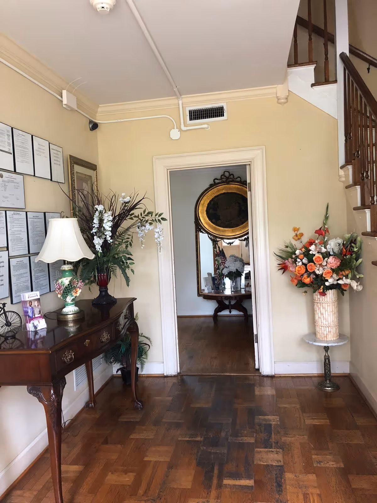 Interior view of a hallway in an assisted living facility with a wooden floor, a wooden table on the left holding a decorative lamp, a flower arrangement, and framed documents on the wall. On the right, there is a tall floral arrangement on a small round table next to a staircase. A doorway leads to another room with a large ornate mirror and a table with flowers.