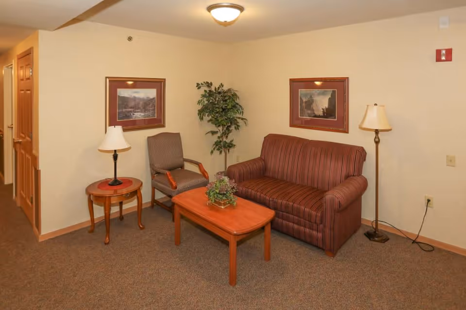Small living room area with a striped loveseat, armchair, coffee table, side table with lamps, and framed art on beige walls.