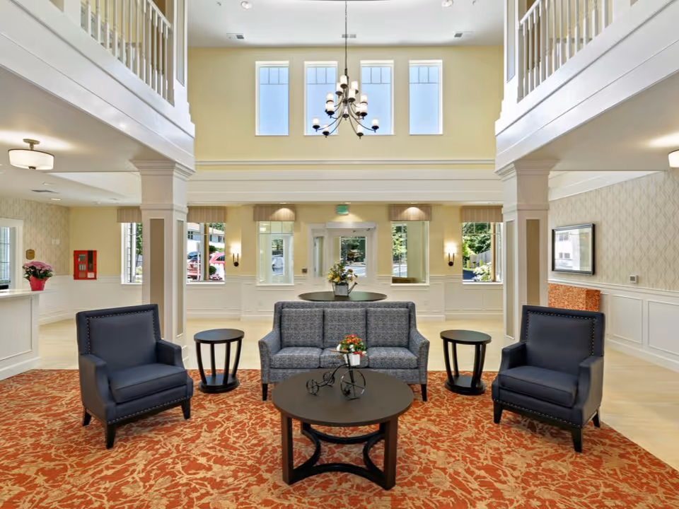 A bright and spacious senior living facility lobby with high ceilings and large windows. The seating area includes a gray sofa, two dark blue armchairs, and three round wooden tables on a red patterned carpet. The walls are light yellow with white wainscoting, and a chandelier hangs from the ceiling. There are columns and a balcony railing visible on the upper level.