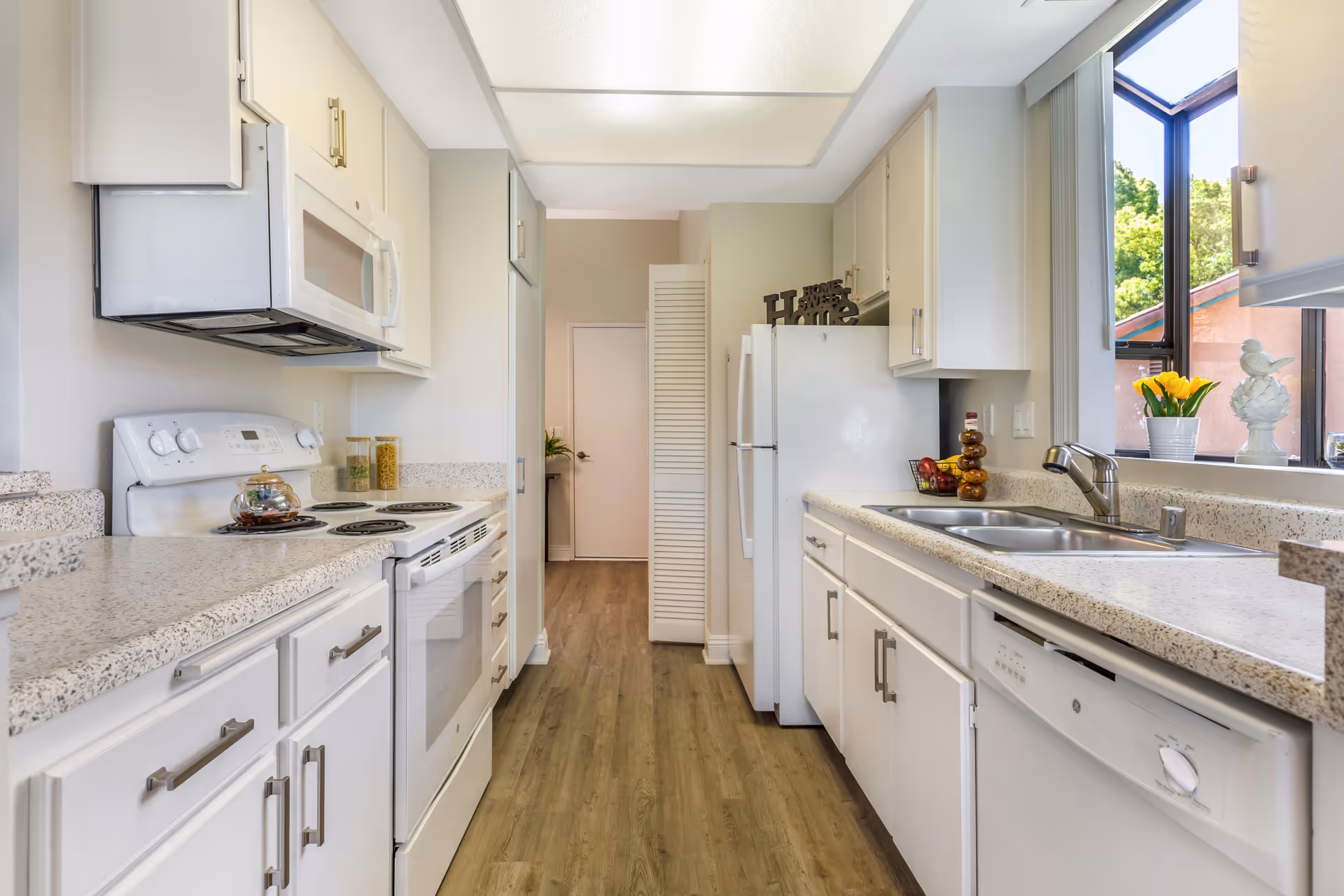 Bright and clean kitchen with white cabinets and appliances including a stove, microwave, refrigerator, and dishwasher. The kitchen has a speckled countertop, a double sink under a window with yellow flowers and a decorative bird, and wood flooring. There is a door at the end of the kitchen and a decorative sign on top of the refrigerator that says 'Home Sweet Home'.