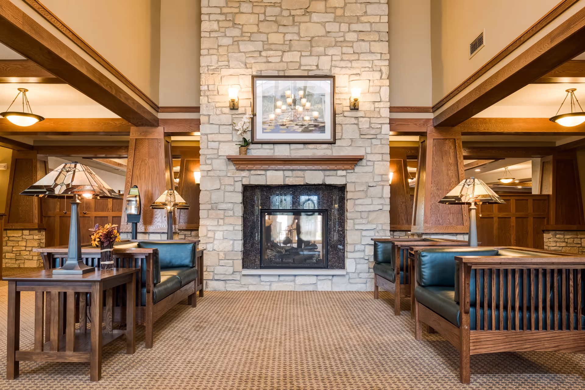 A cozy senior living facility common area featuring a stone fireplace with a framed picture above it. The room has wooden furniture including armchairs with dark green cushions and side tables with decorative lamps. The space is warmly lit with ceiling lights and wall sconces, and has a patterned carpet floor.