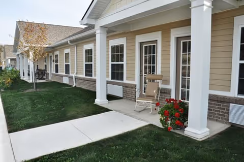 Exterior view of a senior living facility showing a covered walkway with white columns, beige siding, multiple windows, and doors. A rocking chair and a pot of red flowers are placed on the porch area. There is a concrete pathway and green grass lawn in front.