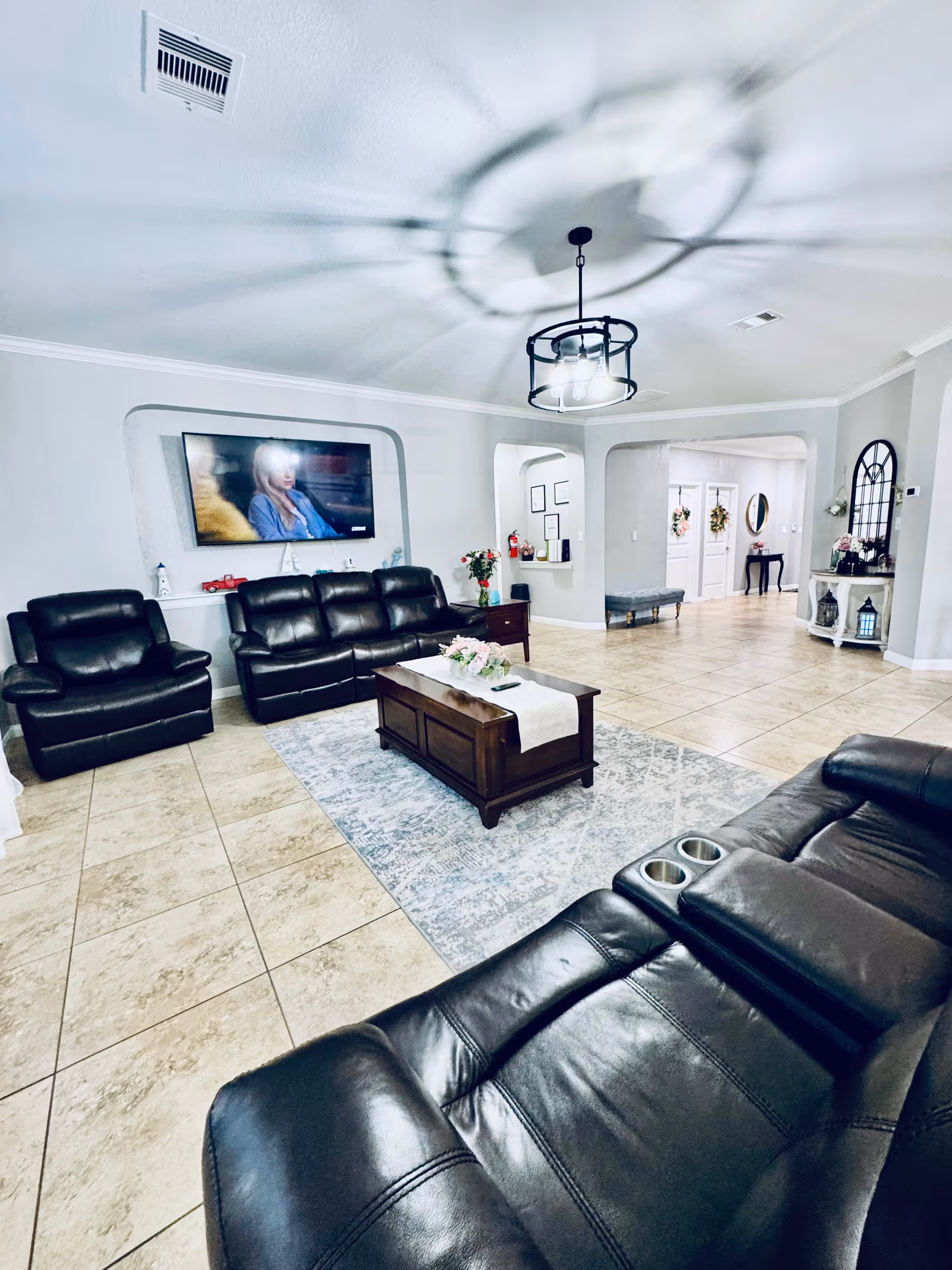 A spacious living room with beige tiled floors and light gray walls. The room features three black leather recliner sofas arranged around a wooden coffee table with a floral centerpiece on a light blue patterned rug. A flat-screen TV is mounted on the wall above the sofas. The ceiling has a modern black light fixture casting shadows, and there are decorative elements such as flowers, a mirror, and framed pictures in the background hallway.