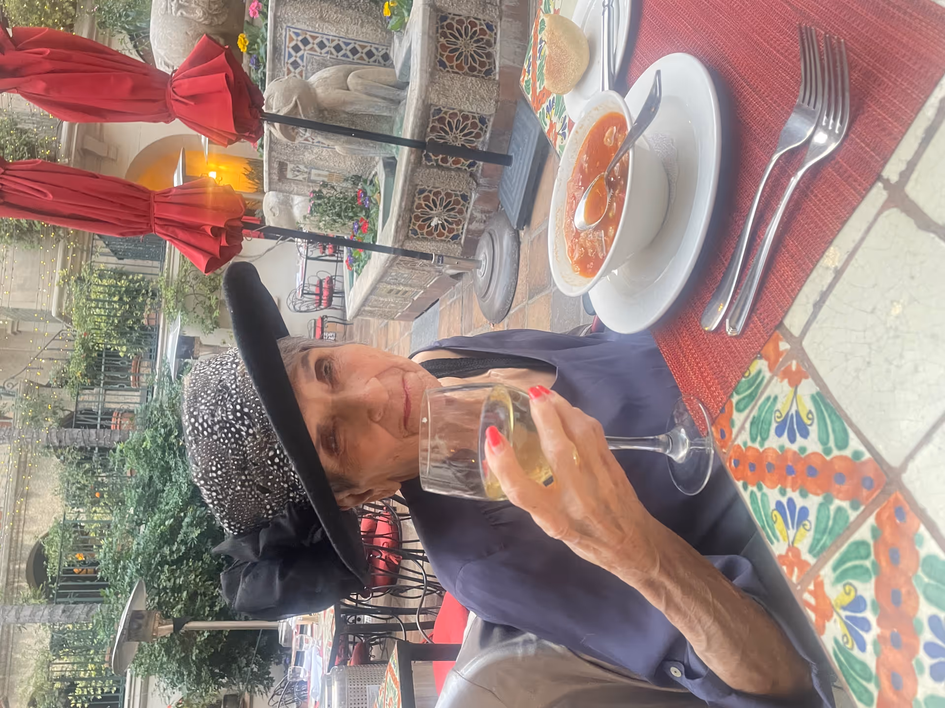 Elderly woman wearing a hat sits at a tiled outdoor restaurant table holding a glass of wine with a bowl of soup and utensils nearby.
