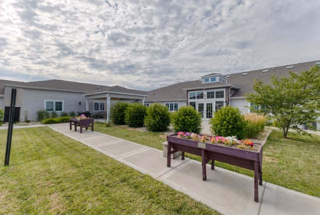 Exterior view of a senior living facility with a paved walkway, green lawn, bushes, and two raised garden beds filled with colorful flowers under a partly cloudy sky.