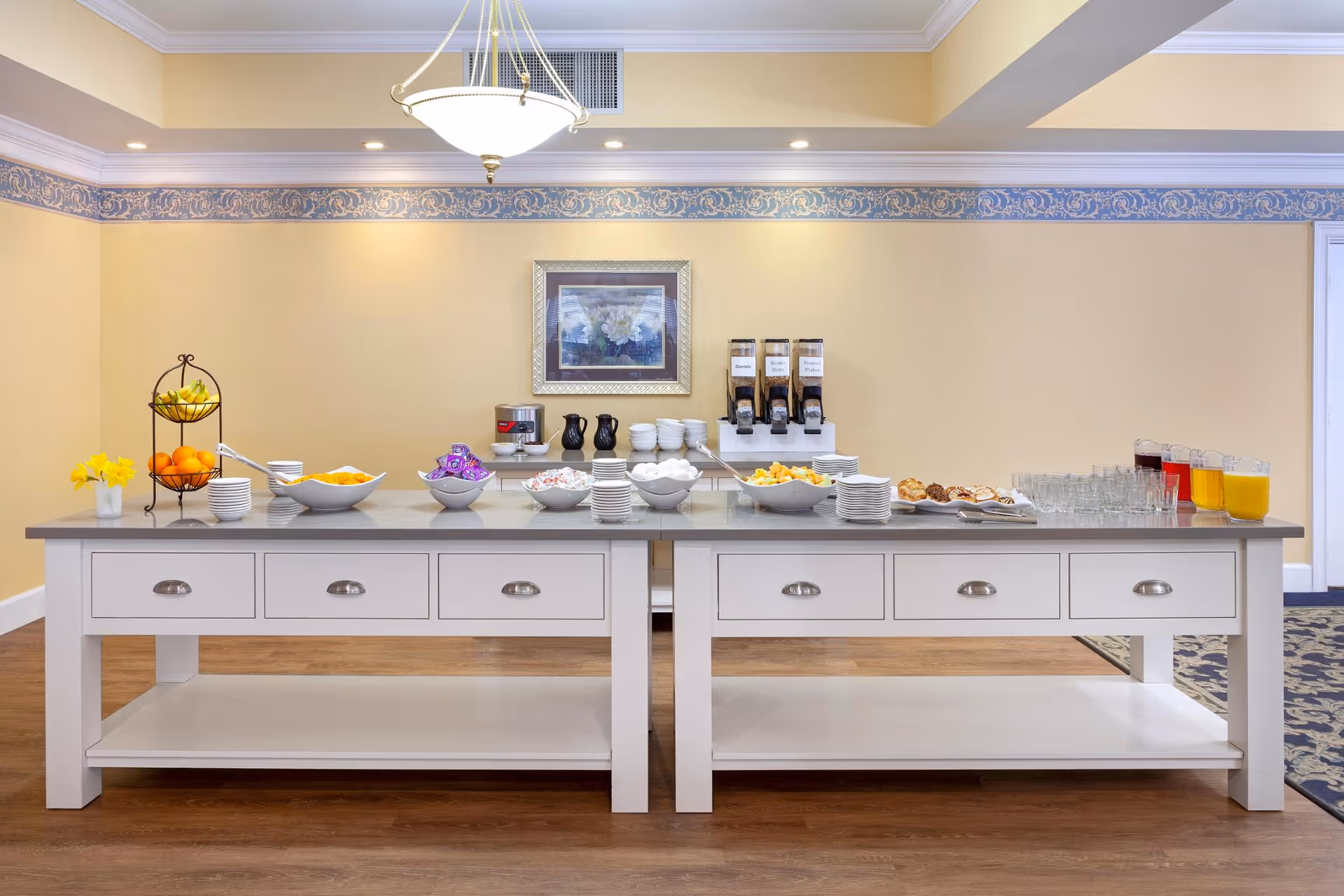 Buffet-style breakfast spread on two white sideboards with bowls of fruit, pastries, drinks and stacked plates in a dining room.