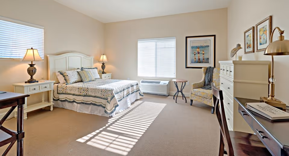 A bright and cozy bedroom with a large bed covered in a patterned quilt and multiple pillows. There are two bedside tables with lamps on either side of the bed. The room has two windows with blinds allowing sunlight to cast shadows on the carpeted floor. A cushioned armchair with a throw blanket is placed near a small round table. A white dresser and a dark wooden desk with a chair and a brass desk lamp are also visible. The walls are decorated with framed artwork.