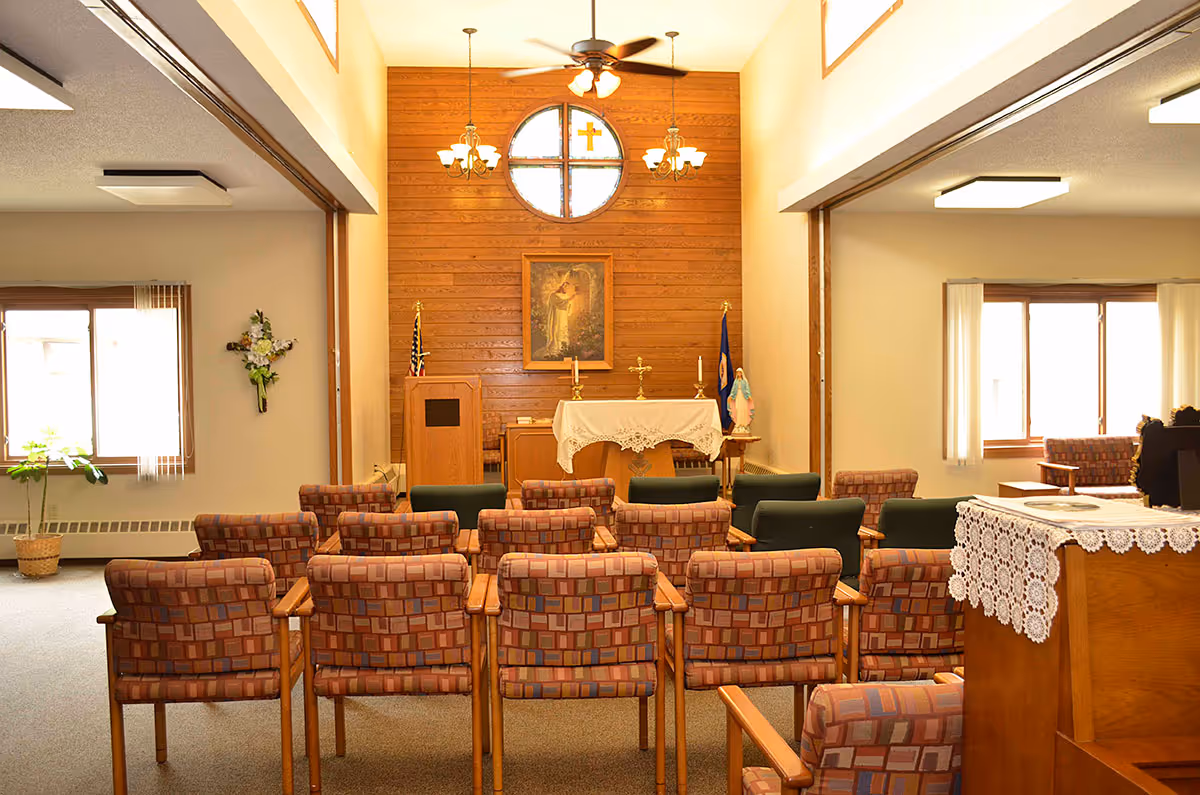 Chapel-like interior with rows of patterned chairs facing an altar against a wood-paneled wall topped by a circular window and cross.