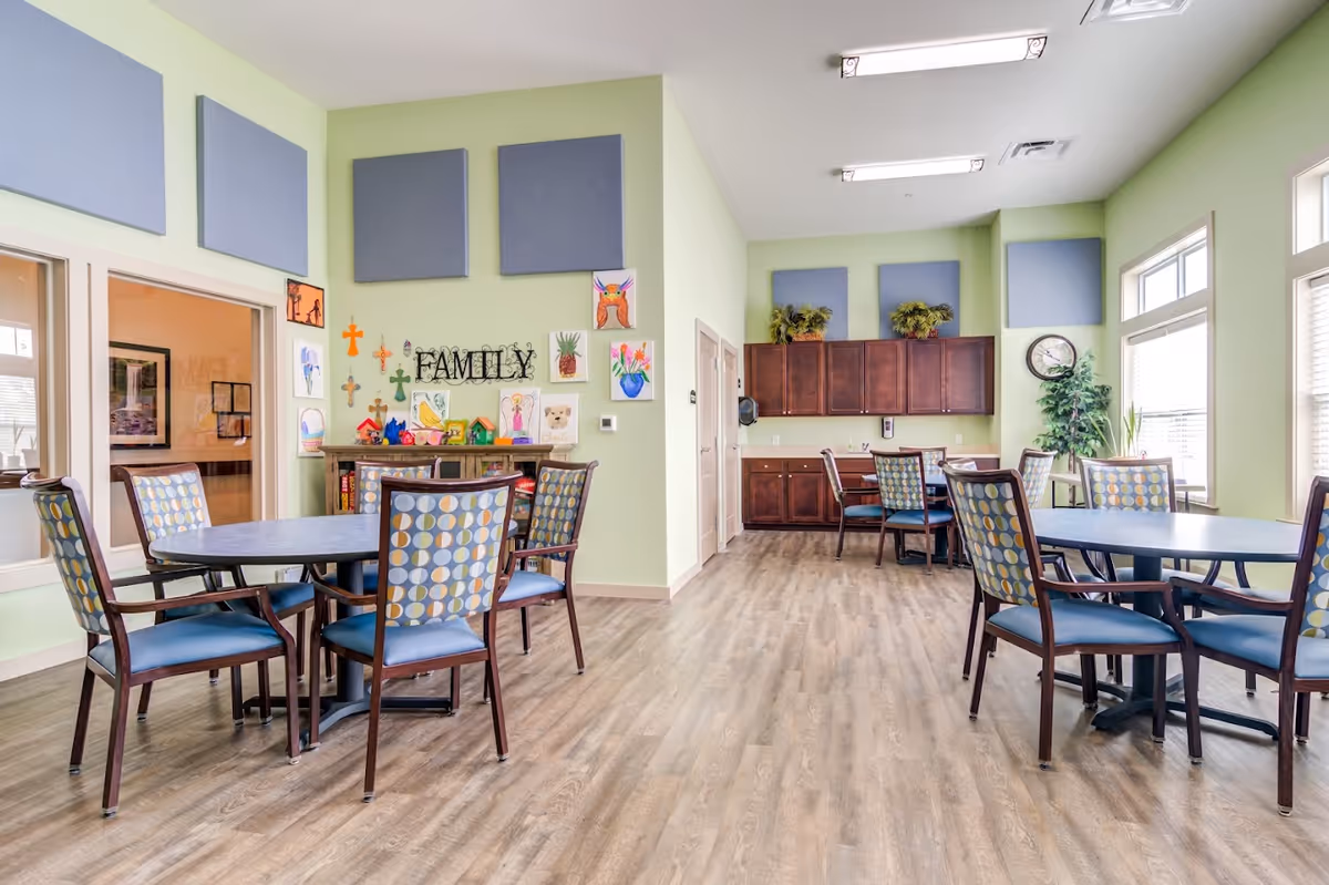 A bright and spacious common area in a senior living facility with light green walls, wooden flooring, and several round tables surrounded by chairs with patterned upholstery. The walls are decorated with colorful artwork and crosses, and there is a cabinet with the word 'FAMILY' displayed above it. Large windows allow natural light to fill the room, and there are plants and a clock on the far wall.