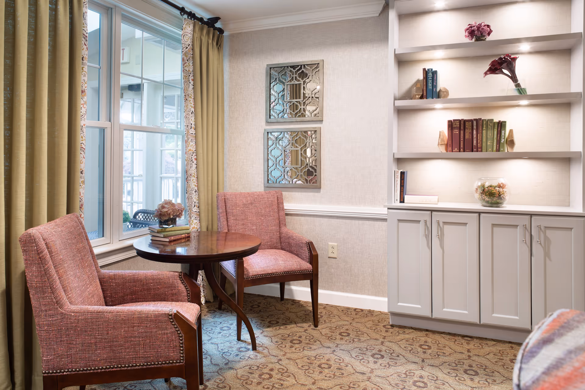 A cozy sitting area with two upholstered pink armchairs around a small round wooden table. The table has a small stack of books and a flower arrangement. Behind the chairs is a large window with beige curtains. On the right side, there is a built-in white cabinet with open shelves displaying books, a flower vase, and decorative items. The walls are light-colored with two decorative mirrors hanging above the chairs.