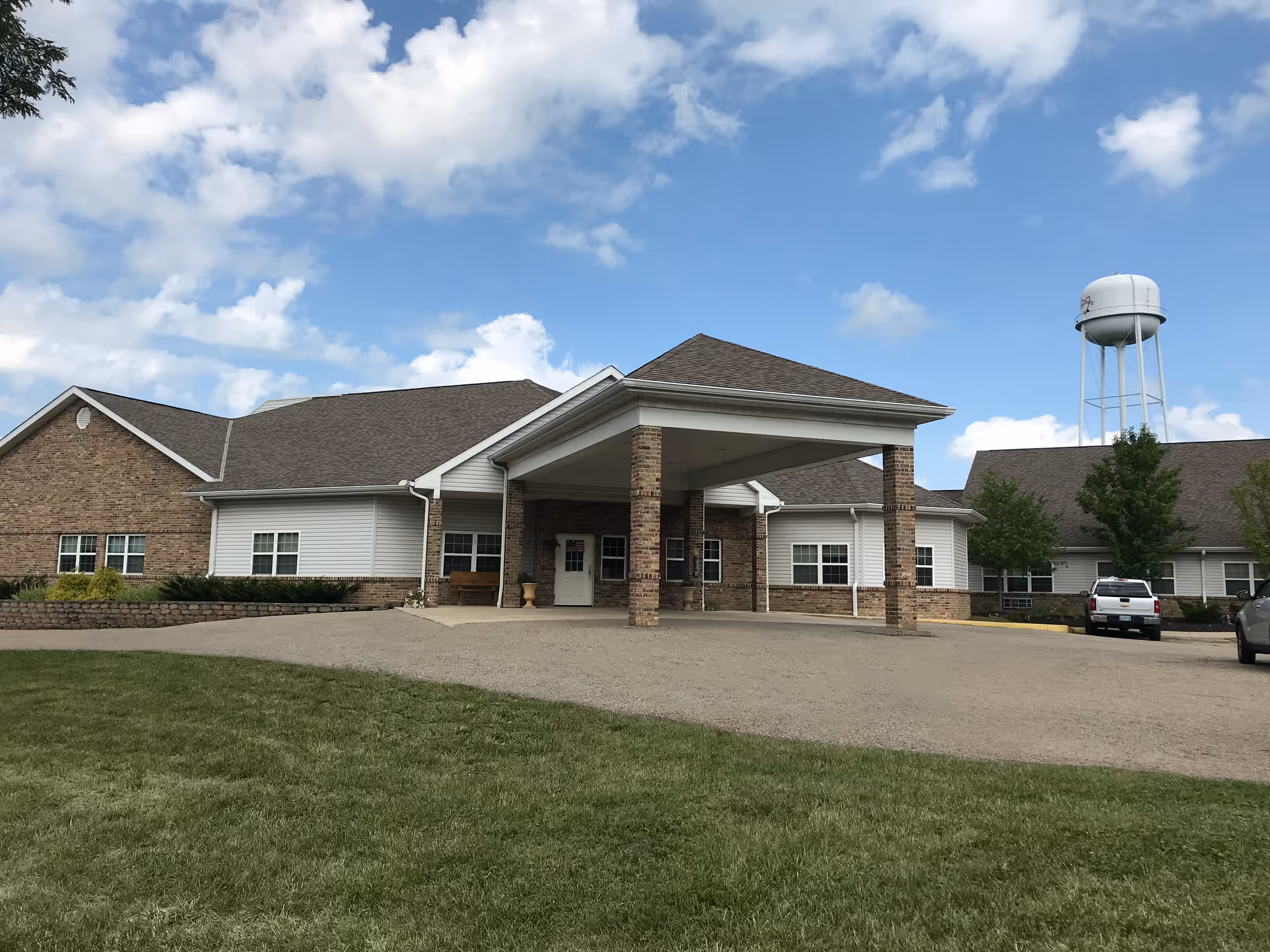 Exterior front entrance of a low-rise brick senior living building with a covered porte-cochere and a water tower in the background.