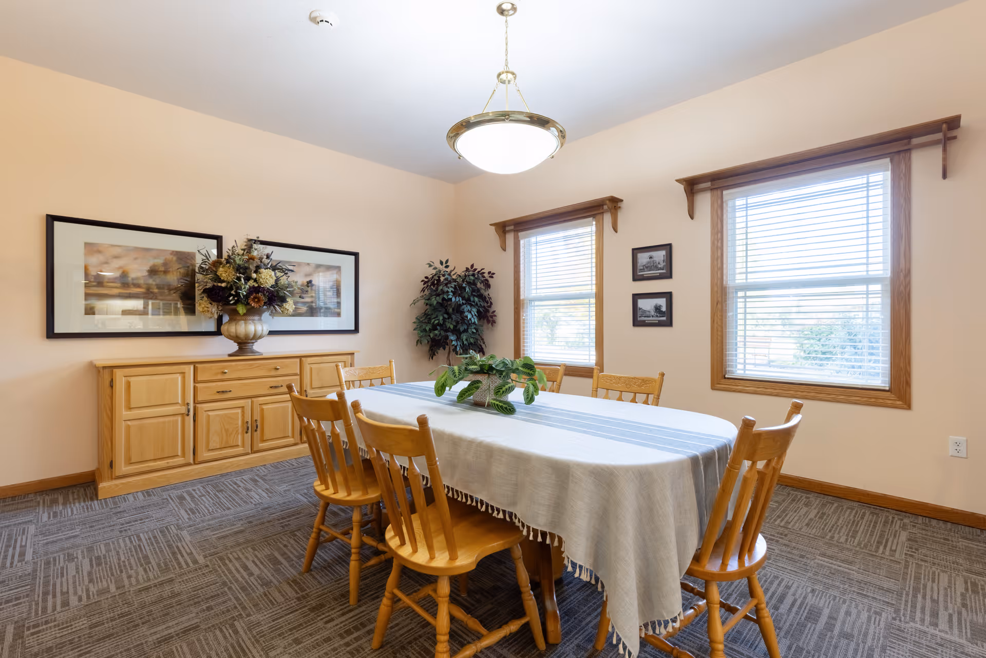 Bright dining room with a rectangular table covered by a tablecloth, wooden chairs, a sideboard with a floral arrangement, and two windows.
