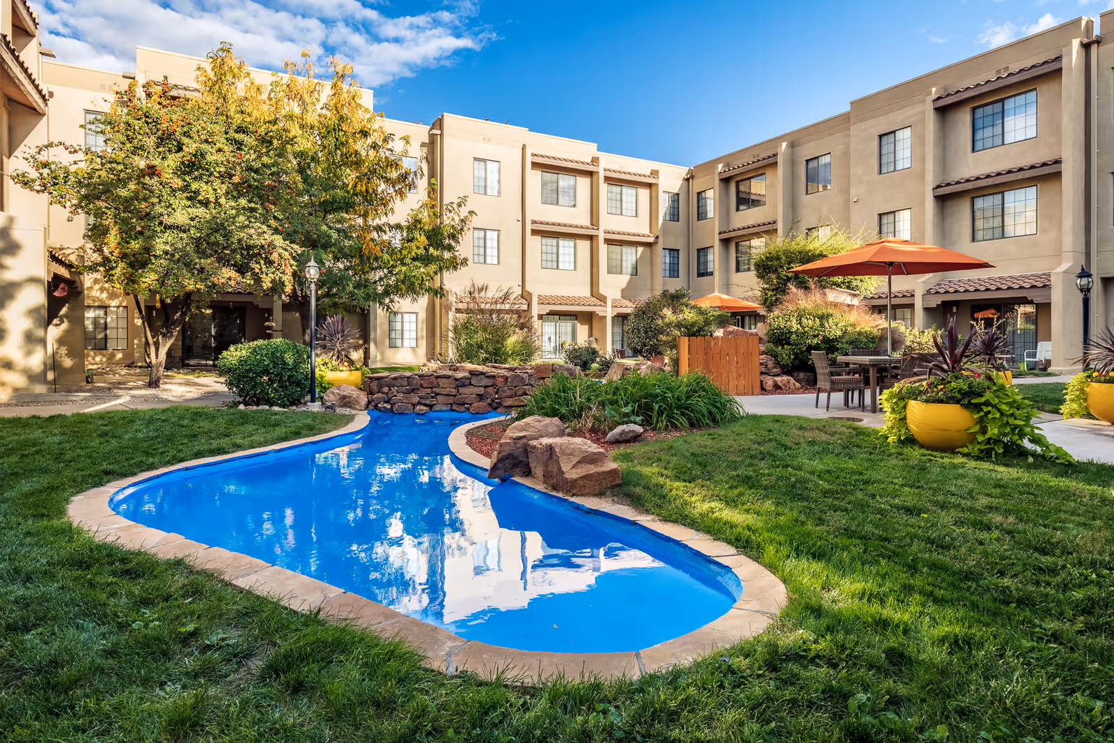 Outdoor courtyard area of a senior living facility with a small blue pond surrounded by grass and landscaping. There are trees, shrubs, and large yellow planters with plants. Several patio tables with orange umbrellas are set up on a paved area near the building. The building is a three-story beige structure with multiple windows and a clear blue sky overhead.