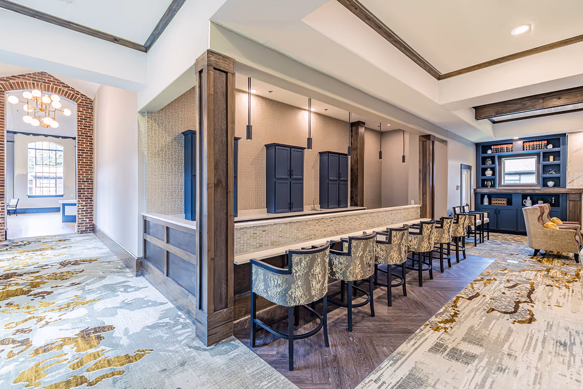 Interior view of a senior living facility lounge area featuring a long counter with eight upholstered bar stools, wooden beams, and pendant lights above. The space includes a carpet with abstract patterns, a brick archway leading to another room with large windows, and built-in blue shelving with decorative items.