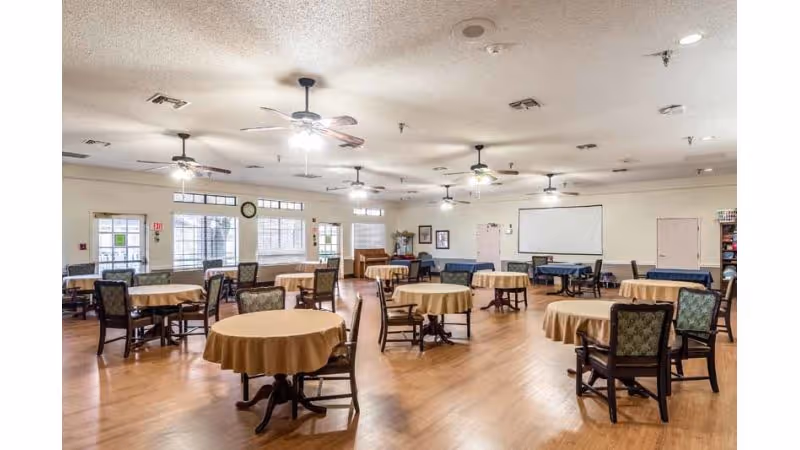 A spacious dining room with multiple round tables covered with beige tablecloths, each surrounded by several chairs. The room has wooden flooring, ceiling fans with lights, large windows letting in natural light, and a projector screen on one wall.