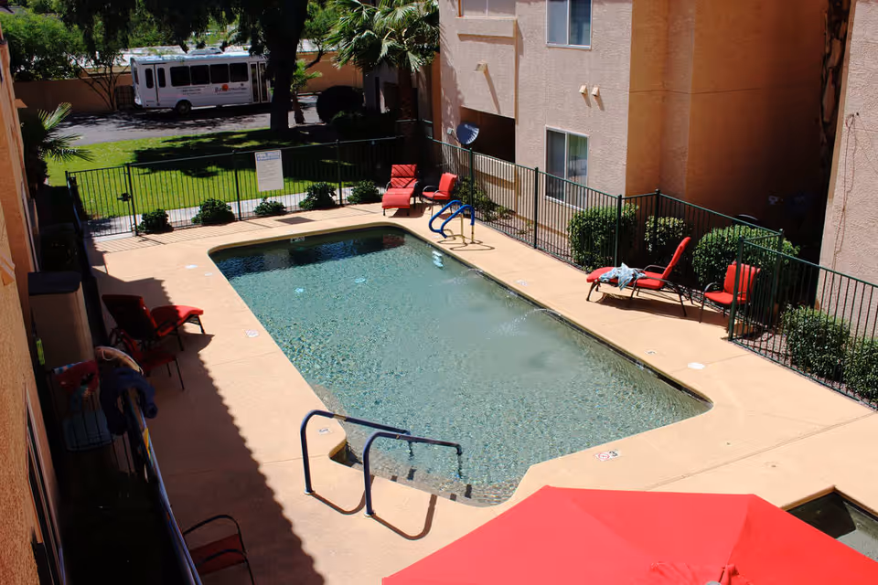 Outdoor swimming pool area at a residential facility with clear water, surrounded by a beige concrete deck. Several red lounge chairs and a red umbrella are placed around the pool. The pool area is enclosed by a black metal fence, with bushes and a grassy area visible beyond the fence. A white shuttle bus is parked in the background near trees and a building.