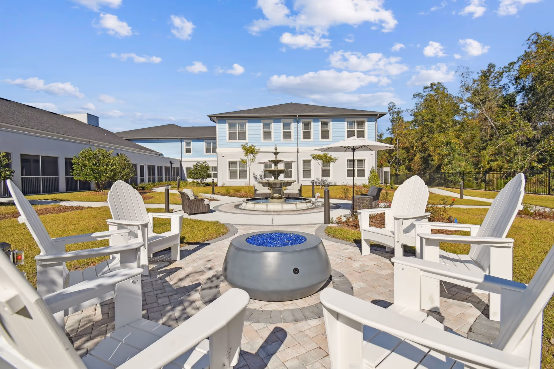 Outdoor courtyard with white Adirondack chairs arranged around a blue-glass fire pit and a fountain in front of a two-story building.