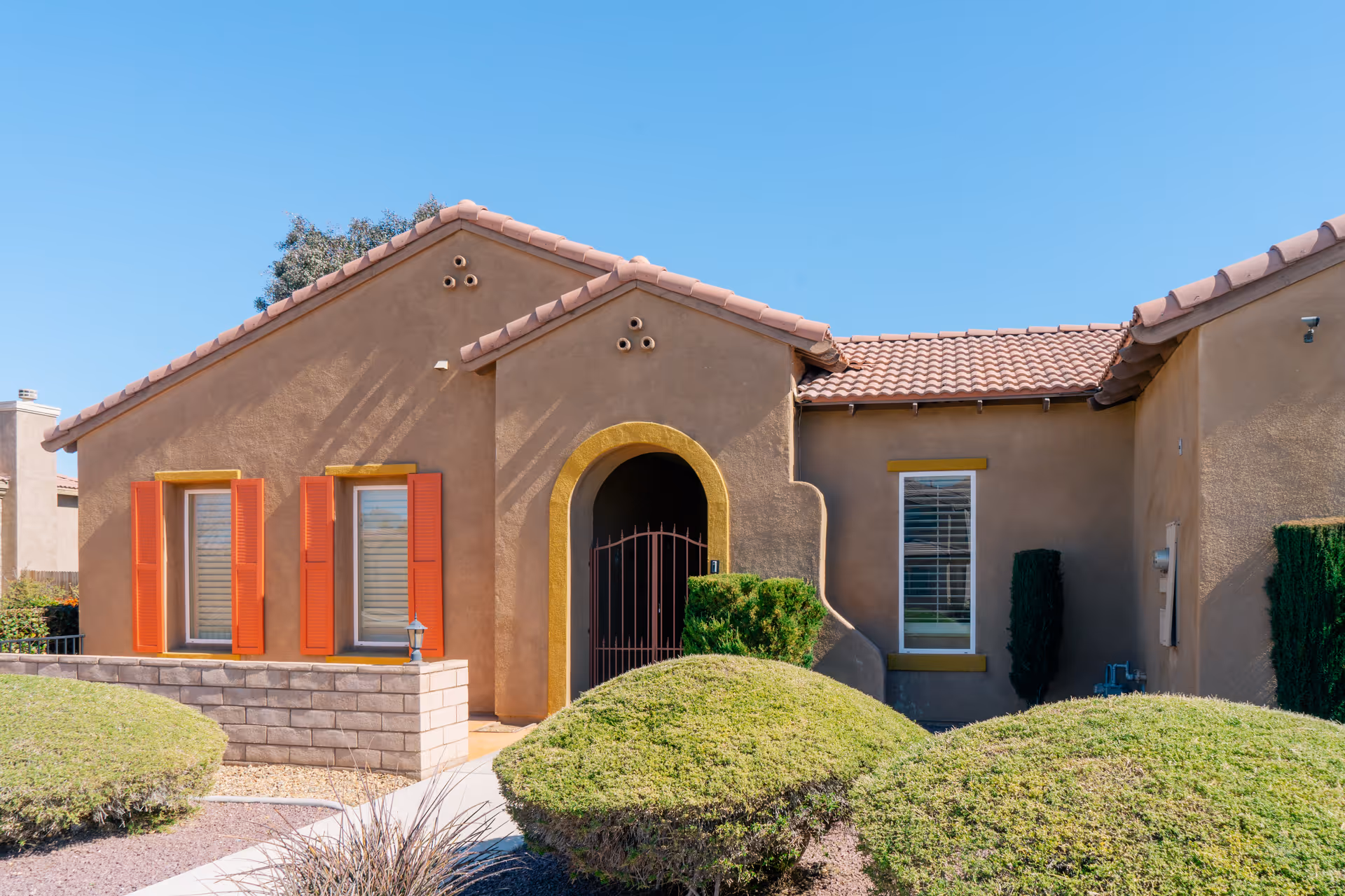 Exterior view of a single-story building with a stucco finish and a tiled roof under a clear blue sky. The building features an arched entrance with a metal gate, two windows with orange shutters on the left, and one window with white blinds on the right. The front yard has neatly trimmed bushes and a paved walkway leading to the entrance.