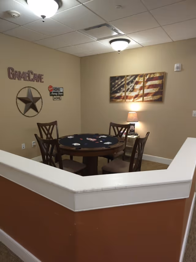 A cozy game room corner with a round poker table surrounded by four wooden chairs. The table has playing cards and poker chips on it. The walls are decorated with a large star, a sign that says 'Game Cave' and 'Sweet Home Away from Home,' and a three-panel American flag artwork. A small lamp on a side table provides warm lighting.