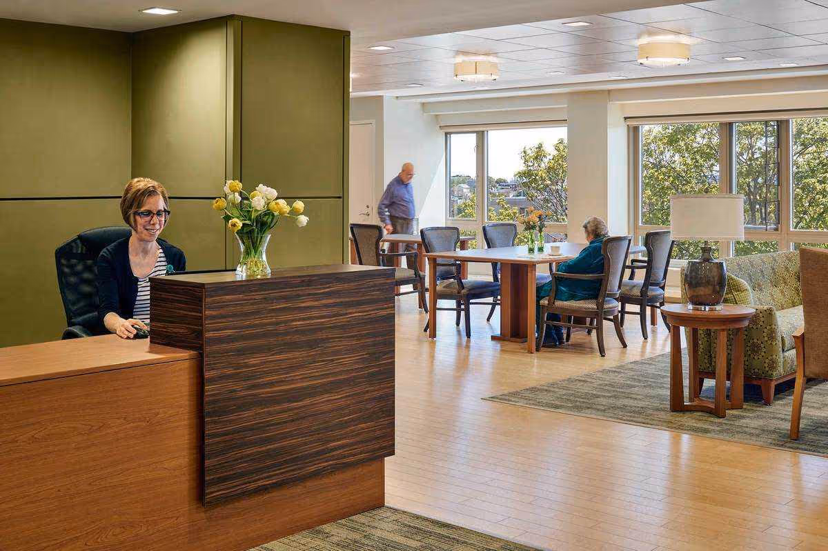A reception area in a senior living facility with a woman sitting behind a wooden desk with a vase of yellow and white flowers. In the background, there are large windows letting in natural light, a man walking, and an elderly woman seated at a table with chairs. There is also a sitting area with a green patterned sofa, a wooden side table, and a lamp.
