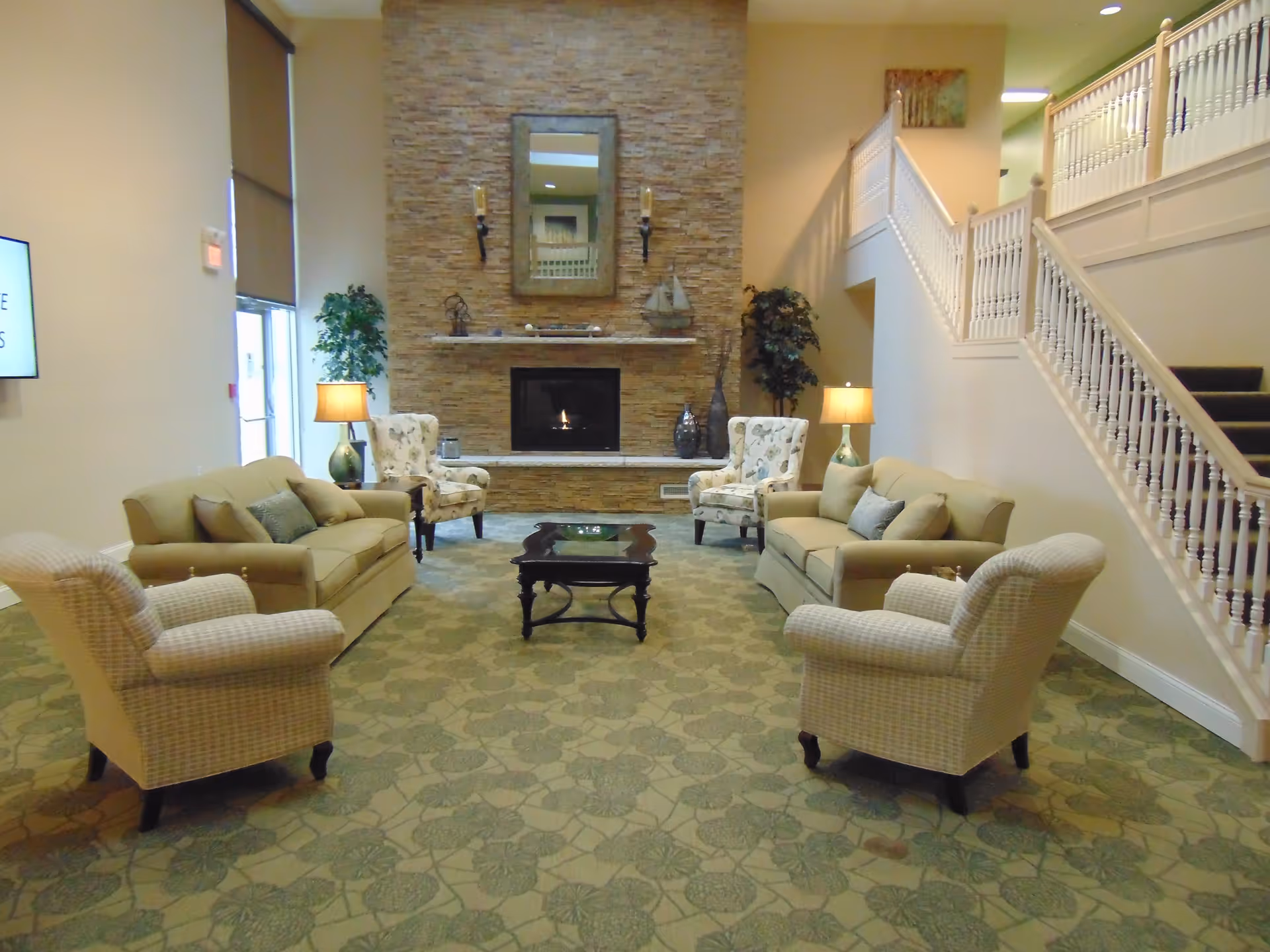 A cozy living room area with two beige sofas, two patterned armchairs, a dark wooden coffee table, and two table lamps. The room features a stone fireplace with a mirror and decorative items above it. There is a staircase with white railings on the right side and large windows on the left side letting in natural light.