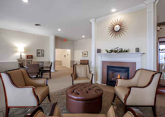 A cozy living room area with beige upholstered armchairs arranged around a round brown ottoman. There is a fireplace with a decorative sunburst mirror above it on the far wall. The room has soft lighting and neutral-colored walls and carpet. In the background, there is a small table with chairs and a lamp on a side table.