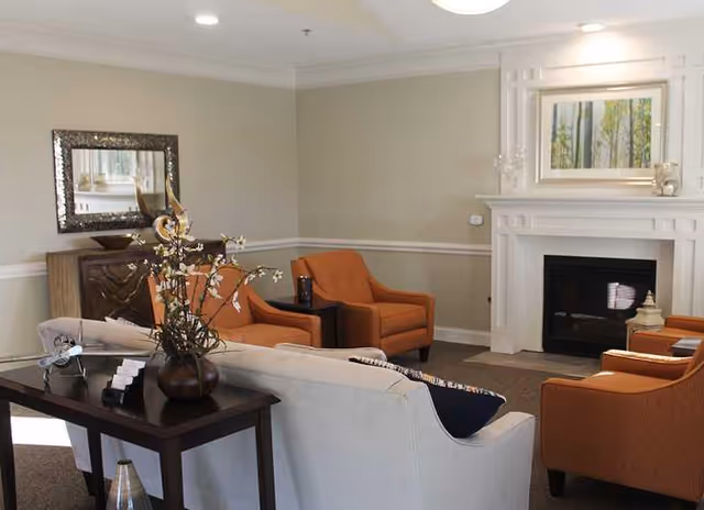 A cozy living room with a beige sofa, four orange armchairs arranged around a white fireplace, a dark wooden console table with a vase of flowers, and a framed mirror on the wall.