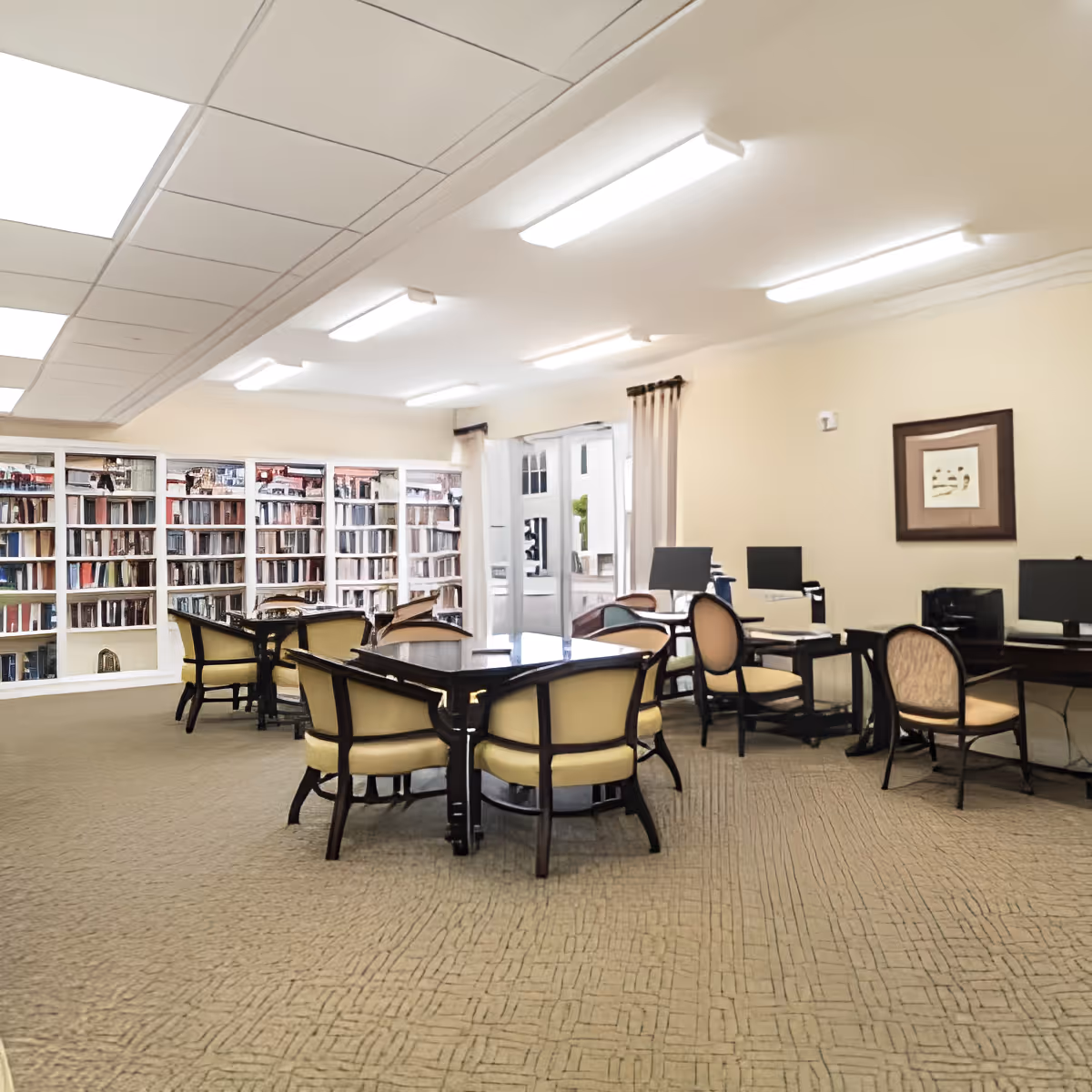 Well-lit communal library/activity room with round tables and upholstered chairs, bookshelves along the back wall and computer stations.