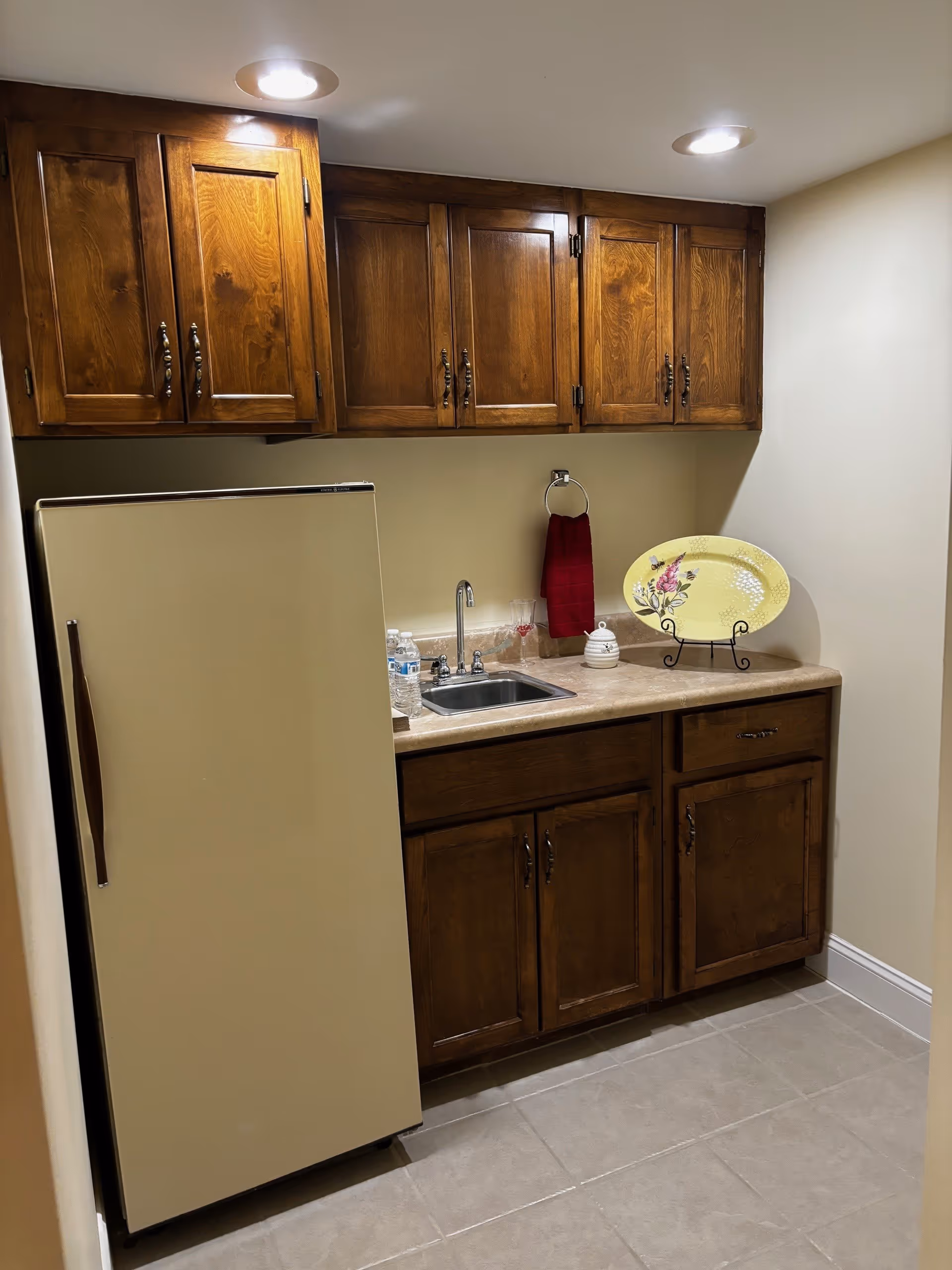 Small kitchen area with wooden upper and lower cabinets, a beige refrigerator, a countertop with a sink, a red hand towel hanging on a ring, a decorative yellow plate on a stand, and two water bottles next to the sink under recessed ceiling lights.