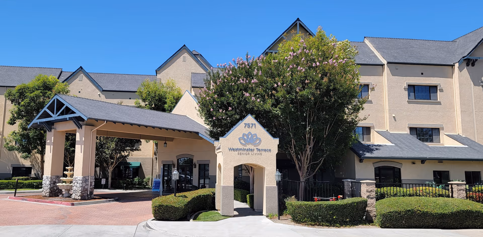 Exterior view of Westminster Terrace Senior Living building with a covered entrance, manicured bushes, trees, and clear blue sky.