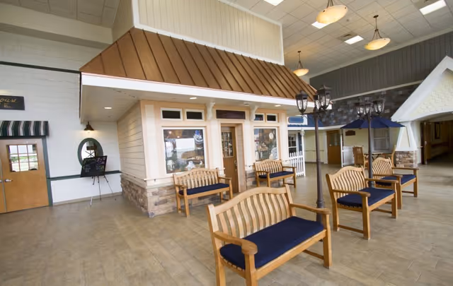 Indoor common area designed to look like a small village street with wooden benches featuring blue cushions, street lamp posts, and storefront-style facades with windows and doors under a high ceiling.