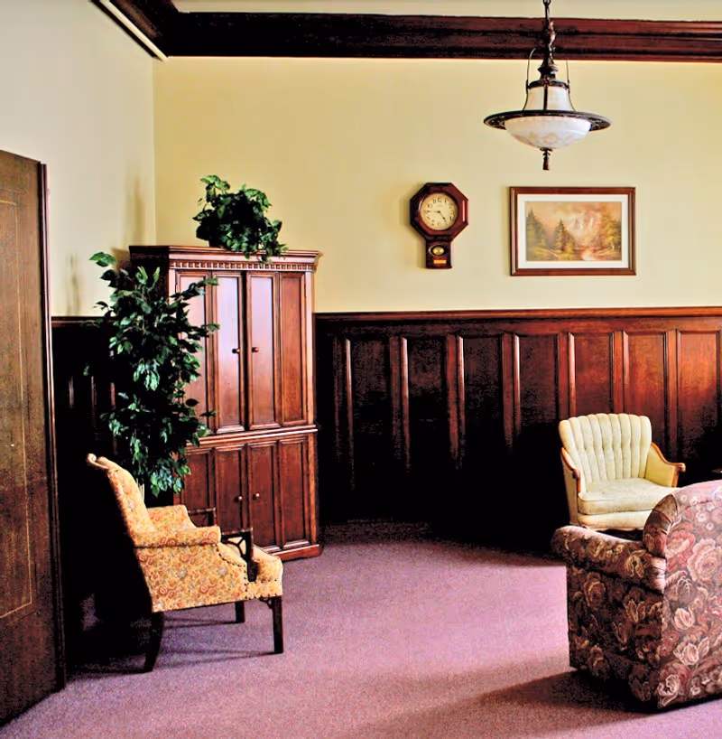 A cozy living room area with vintage-style furniture including a floral armchair, a cream-colored cushioned chair, and a wooden cabinet with a potted plant on top. The walls have dark wood paneling on the lower half and light paint above, decorated with a wall clock and a framed landscape painting. A hanging light fixture is visible on the ceiling.