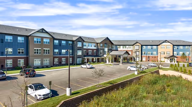 Exterior view of a large three-story senior living facility building with multiple windows and entrances. The building has a mix of beige, blue, and brick red colors. There is a parking lot in front with several parked cars and a landscaped area with grass and small trees. The sky is partly cloudy.