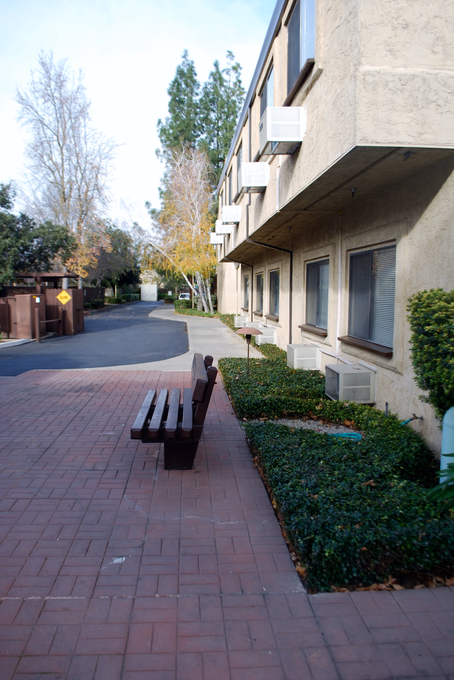 Outdoor view of a beige two-story apartment building with multiple windows and air conditioning units. A paved walkway with a wooden bench runs alongside the building, bordered by green shrubs. Trees and a driveway are visible in the background.