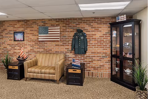 A cozy sitting area with a striped loveseat flanked by two black side tables, one holding a model sailboat and the other a small American flag decoration. Behind the loveseat is a brick wall adorned with a wooden American flag, a framed picture, and a military jacket. To the right is a black display cabinet with glass doors containing various decorative items and small American flags. The floor is carpeted with a patterned design, and there are two potted plants on either side of the scene.