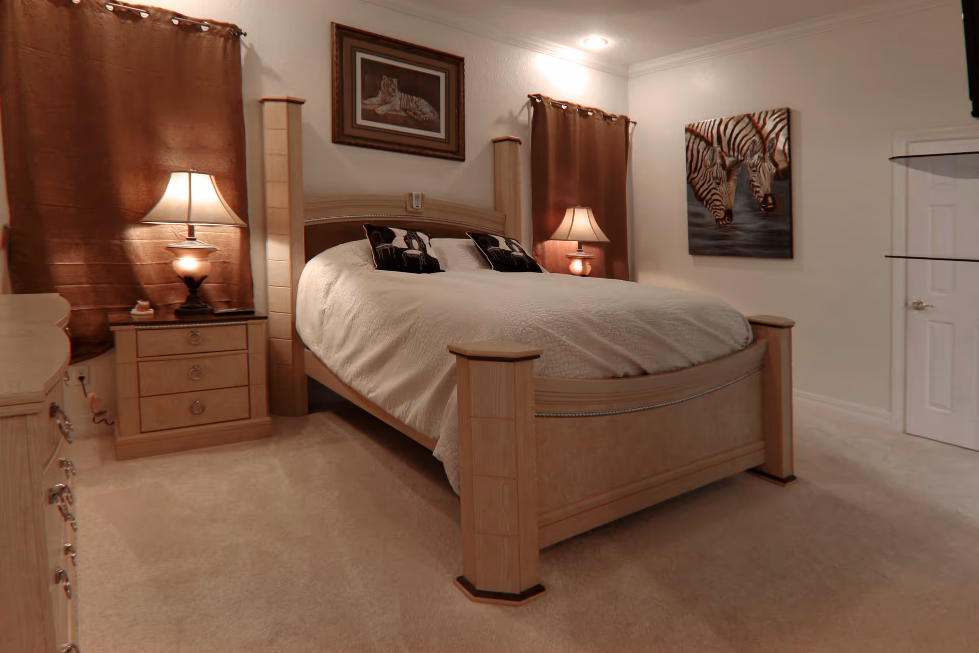 A cozy bedroom with a large wooden bed featuring a white quilt and two decorative pillows. There are two nightstands with lamps on either side of the bed, brown curtains on the windows, and two framed animal paintings on the walls. The room has beige carpeting and white walls.
