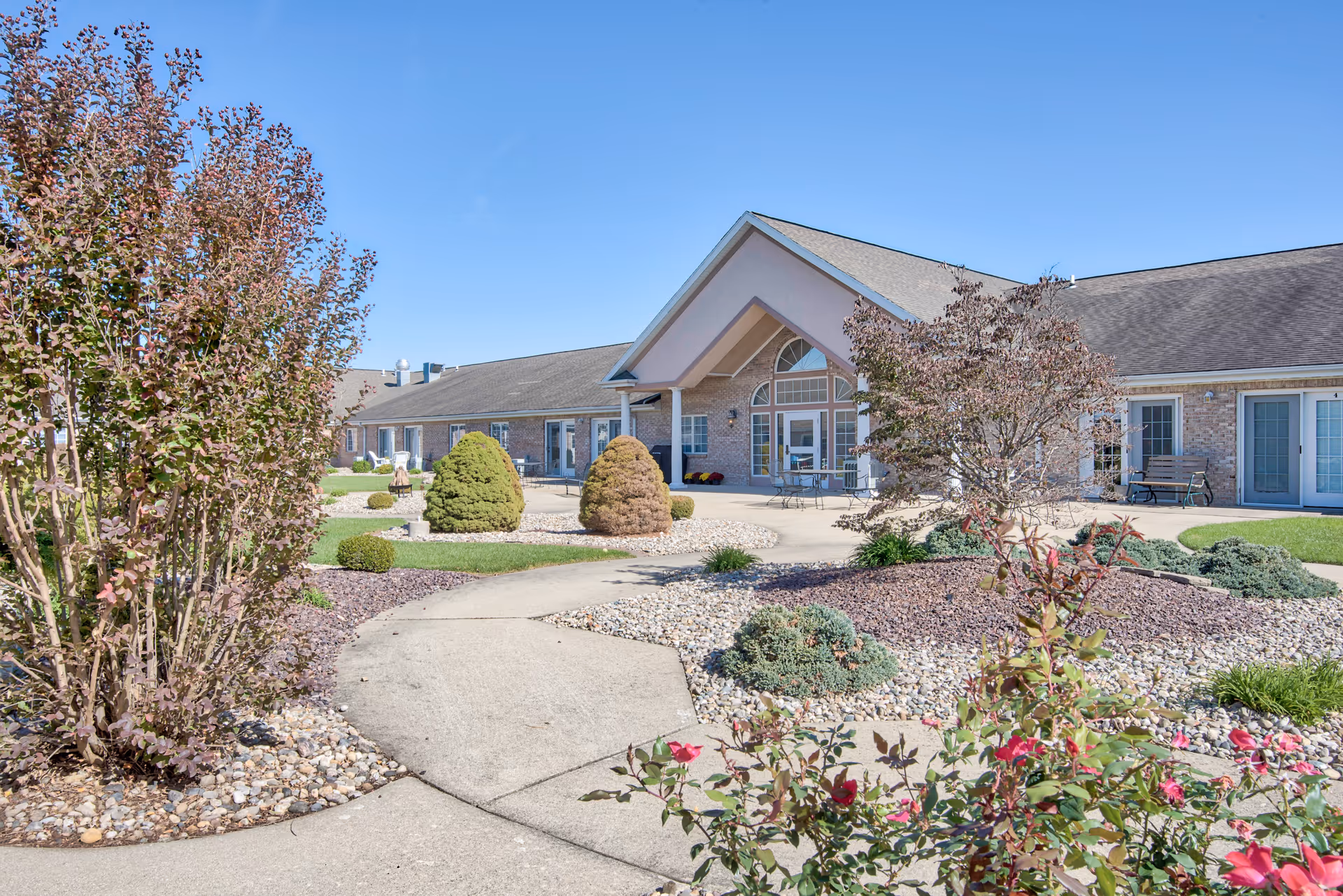Exterior view of The Villas at St. James facility showing a landscaped garden with bushes, small trees, and flowering plants along a curved concrete pathway leading to the building entrance under a peaked roof. The building has brick walls and multiple windows and doors.