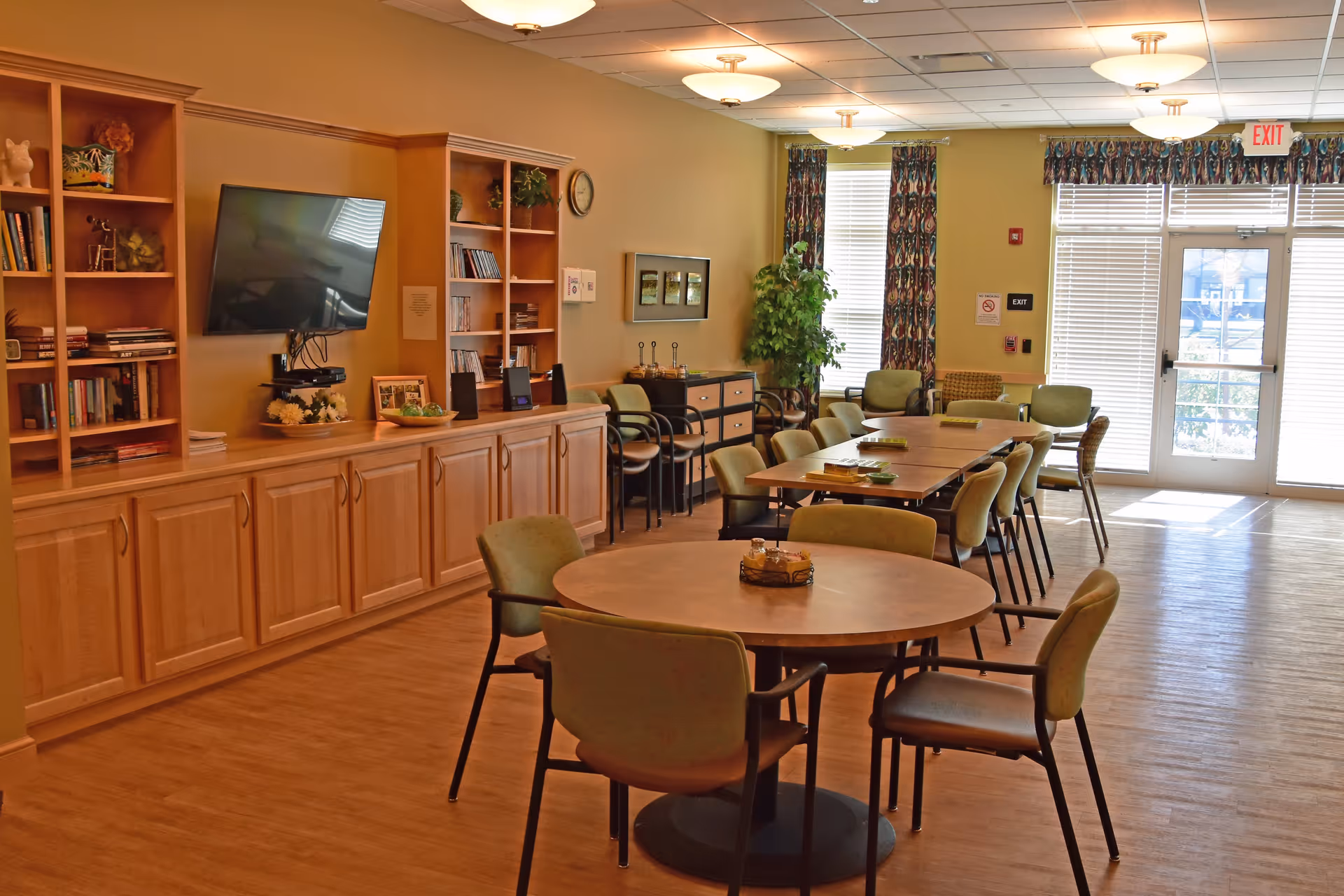 A well-lit common area in an assisted living facility featuring round and rectangular tables with green cushioned chairs. The room has wooden built-in shelves and cabinets along one wall, a mounted flat-screen TV, decorative plants, and large windows with patterned curtains letting in natural light. The floor is wooden, and there are ceiling lights evenly spaced across the room.