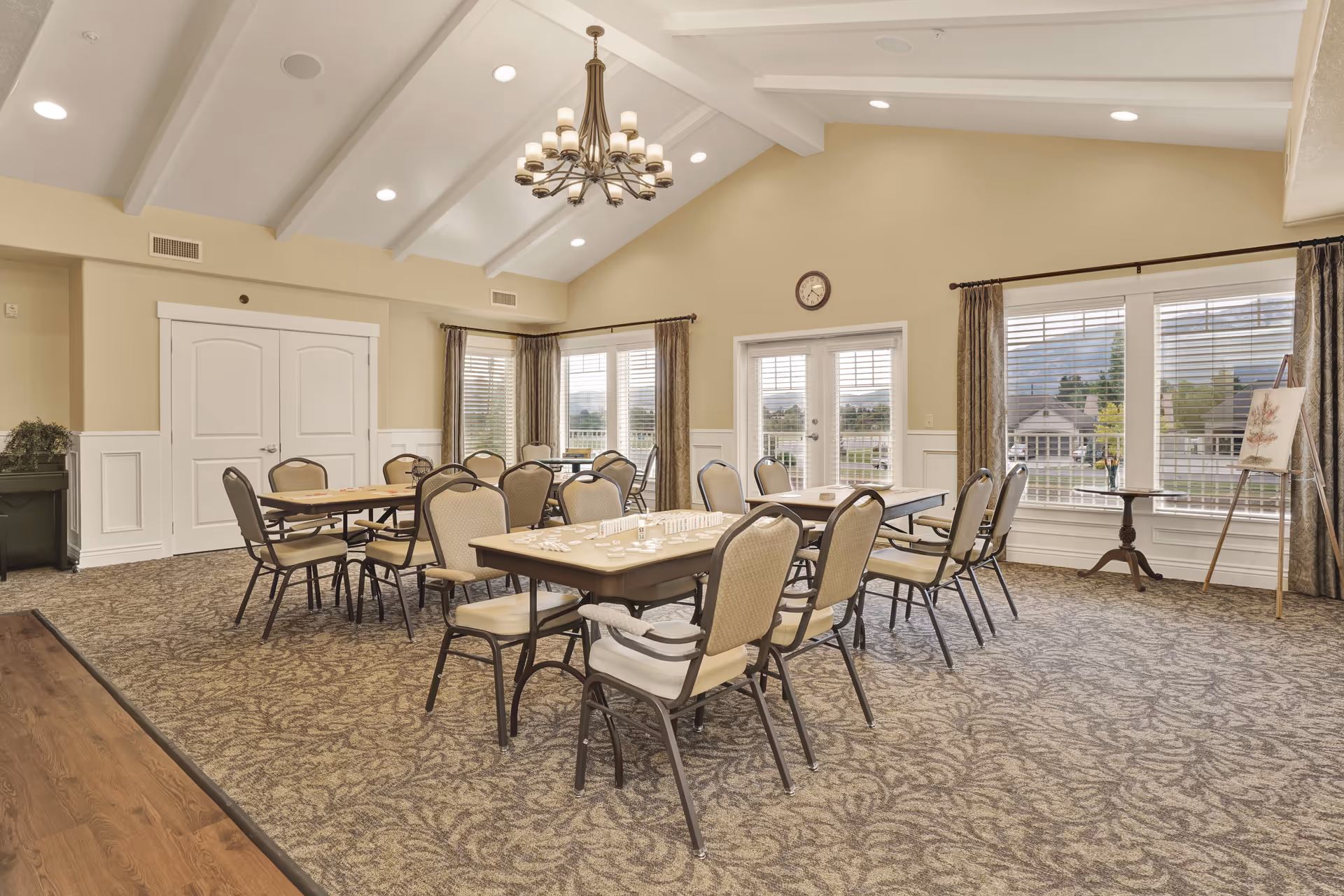 A bright and spacious common room with beige walls and carpeted floor featuring several tables and chairs arranged for group activities. The room has large windows with curtains allowing natural light to enter, a chandelier hanging from the vaulted ceiling, and a clock on the wall above a set of French doors. Some tables have game pieces on them, suggesting the space is used for social or recreational activities.