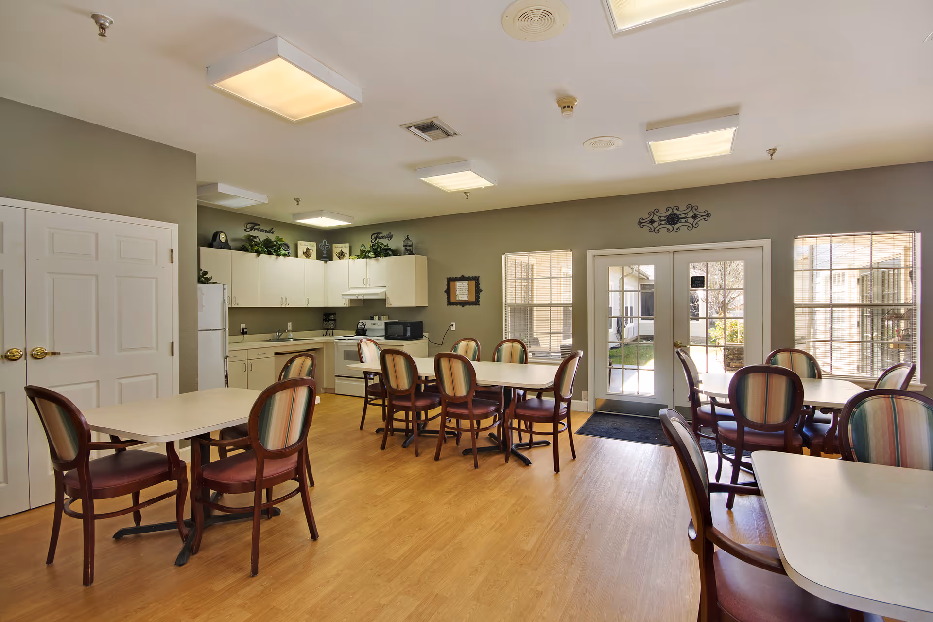 A bright and clean dining area in a senior living facility featuring several tables with chairs arranged around them. The room has light wood flooring, beige walls, and a kitchen area with white cabinets, a refrigerator, microwave, and stove. Large windows and glass doors allow natural light to enter, showing a glimpse of an outdoor garden area.