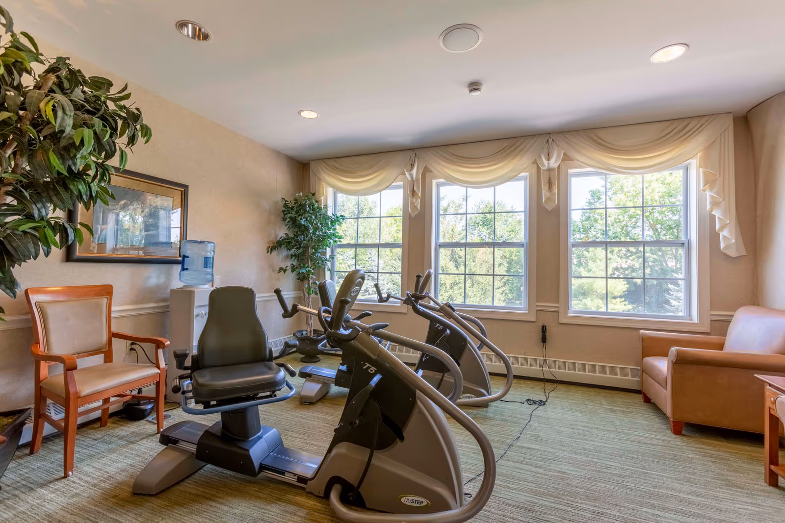 A bright exercise room with large windows letting in natural light, featuring two recumbent exercise bikes, a water cooler, a wooden chair, a leather armchair, and potted plants. The room has beige walls, green carpet, and decorative window valances.