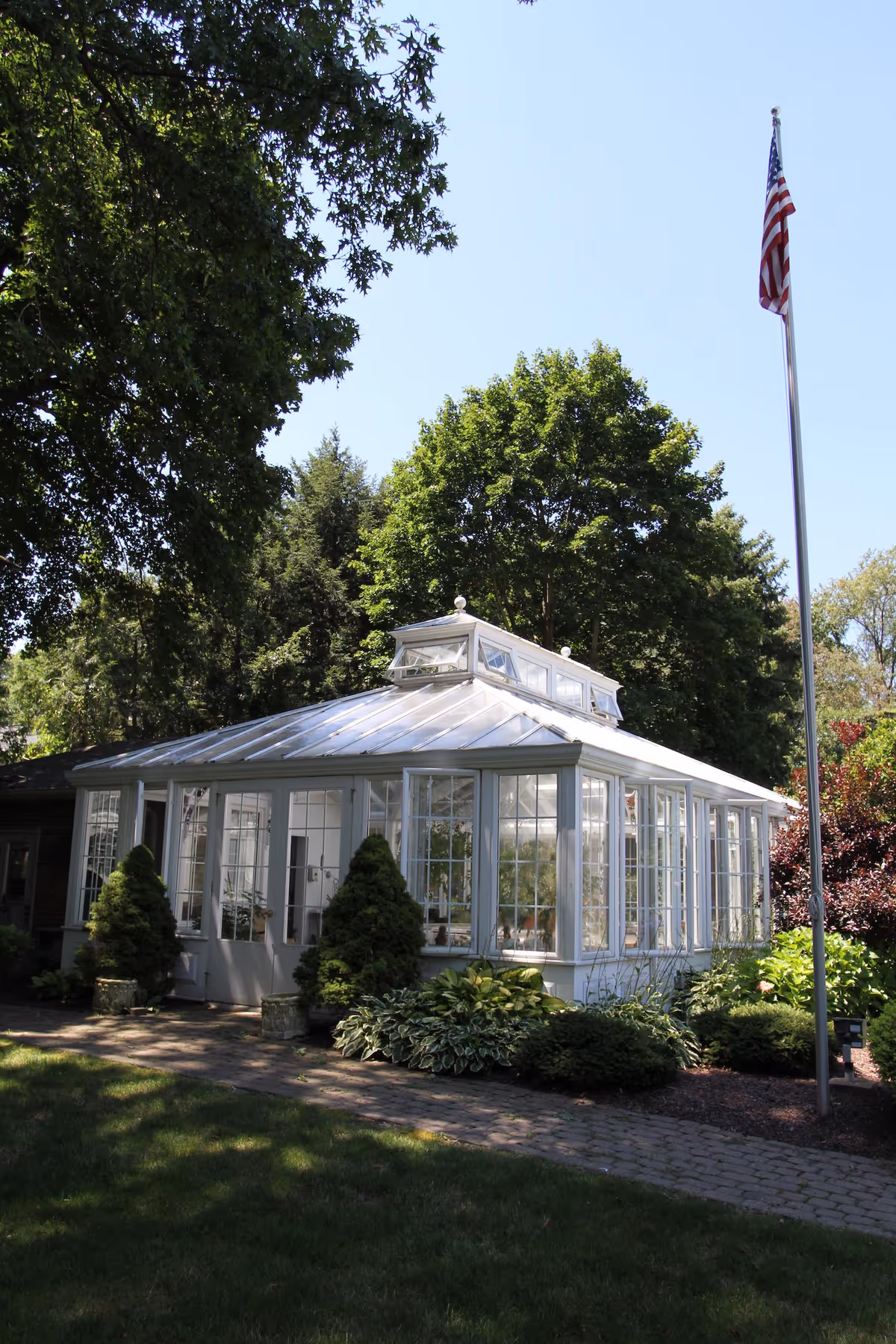 White glass conservatory-style building surrounded by trees and landscaping with an American flag on a pole.
