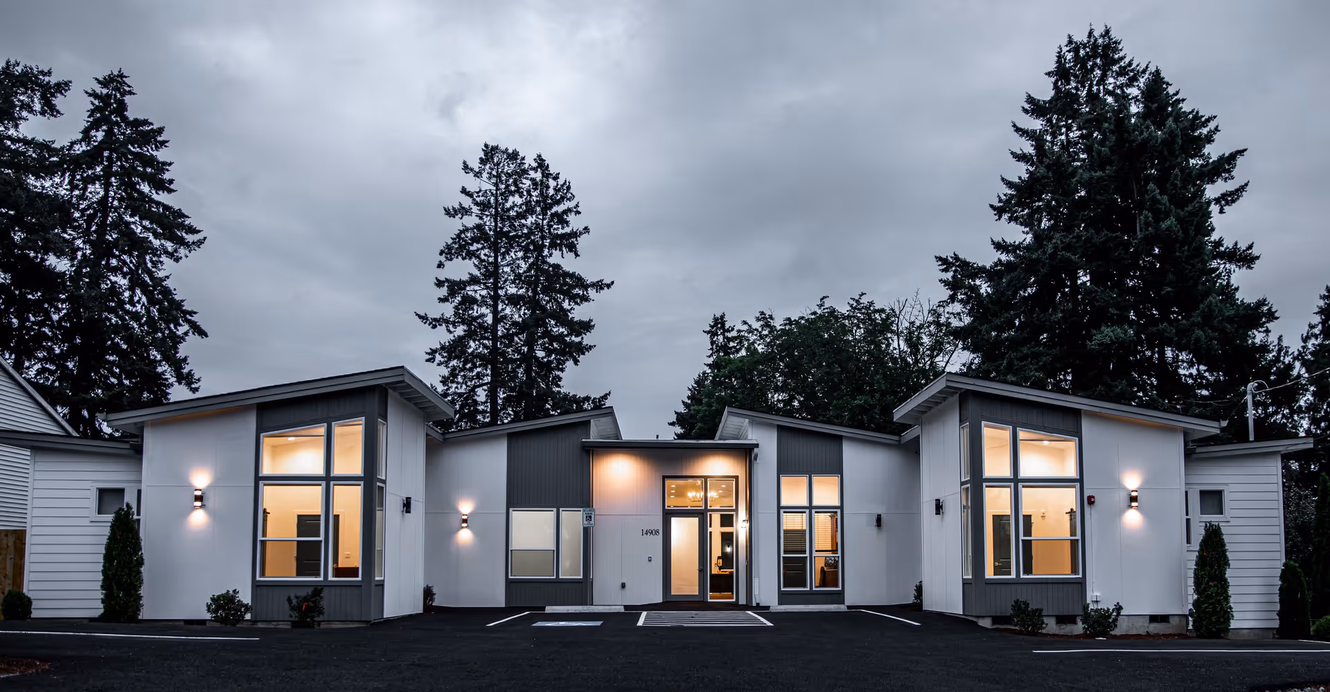 Exterior view of a modern single-story building at dusk with large windows and exterior lights on, surrounded by tall trees and a paved parking area in front.